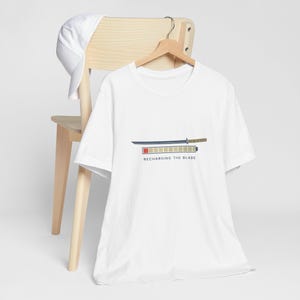 May include: White t-shirt with a graphic of a katana and a battery charging bar, with the text "RECHARGING THE BLADE". A white baseball cap rests on a wooden chair.