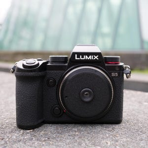 May include: A black Lumix S5 camera with a textured grip and a large lens. The camera has the Lumix logo on top and the model number S5 on the side. The background is blurred, suggesting an outdoor setting.