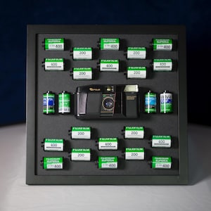 May include: A black and silver film camera with a black lens and a silver button on the top. The camera is surrounded by a variety of film canisters, including Fuji Superia 400, Fuji 200, and Fuji 400 film.