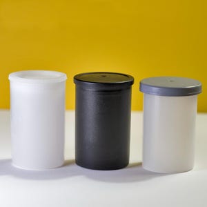 May include: Three film canisters, one white, one black, and one gray. The canisters are made of plastic and have a screw-on lid.