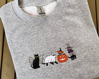 May include: A gray sweatshirt featuring embroidered Halloween-themed cats. The cats are depicted as a bat, a ghost, a pumpkin, and a witch. The sweatshirt is made of a soft material and has a crew neck.