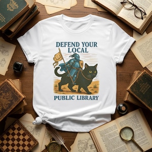 Op de afbeelding: Wit t-shirt met een afbeelding van een ridder op een zwarte kat, met een vlag. De tekst "DEFEND YOUR LOCAL PUBLIC LIBRARY" staat in blauw. Het ontwerp heeft een vintage, middeleeuwse stijl.