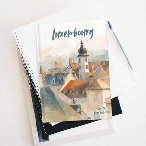 Può includere: Un taccuino con un'illustrazione ad acquerello del paesaggio urbano del Lussemburgo, con un campanile e dei tetti. La parola "Lussemburgo" è scritta in un carattere script. Il taccuino è sopra altri taccuini e una penna.