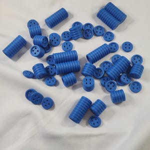 Puede incluir: Una colección de piezas de construcción de juguete de plástico azul, que incluyen componentes cilíndricos y circulares. Los cilindros tienen lados acanalados y las piezas circulares tienen cuatro agujeros. Las piezas están esparcidas sobre una superficie blanca.