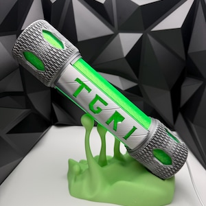May include: A cylindrical object with a silver, textured exterior and a bright green core. The silver casing features the letters TGKI in a cutout design, revealing the green interior. It rests on a light green, organic-shaped stand.