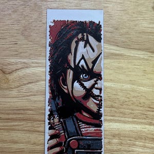 May include: A rectangular bookmark with a Chucky illustration. The image shows Chucky's face, with red hair, blue eyes, and a stitched scar. The word "CHU" is visible. The bookmark is a horror film-themed item.