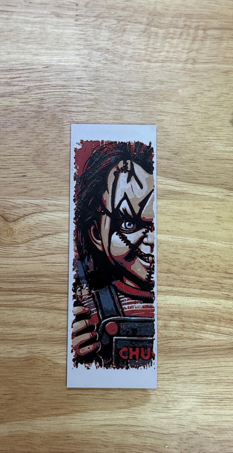 May include: A rectangular bookmark with a Chucky illustration. The image shows Chucky's face, with red hair, blue eyes, and a stitched scar. The word "CHU" is visible. The bookmark is a horror film-themed item.