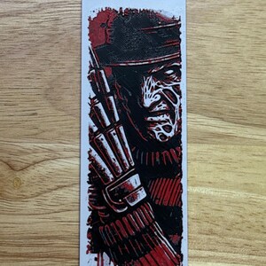 May include: A rectangular bookmark with a red and black illustration of a character wearing a hat and glove. The character's face is white with red lines. The bookmark is on a wooden surface.