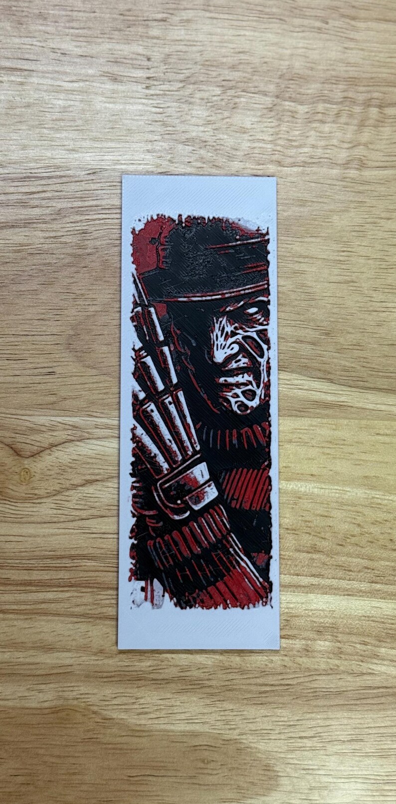 May include: A rectangular bookmark with a red and black illustration of a character wearing a hat and glove. The character's face is white with red lines. The bookmark is on a wooden surface.