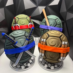 May include: Four circular turtle shells in olive, dark green, and tan, each featuring a colored mask and signature weapon. The shells include red, purple, blue, and orange masks with sai, a bo staff, katanas, and nunchucks mounted on circular stands.