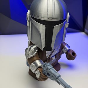 May include: A silver and black Mandalorian action figure, holding a gray blaster pistol. The figure has a detailed helmet and armor, with brown and silver accents. The background is a blue geometric pattern.