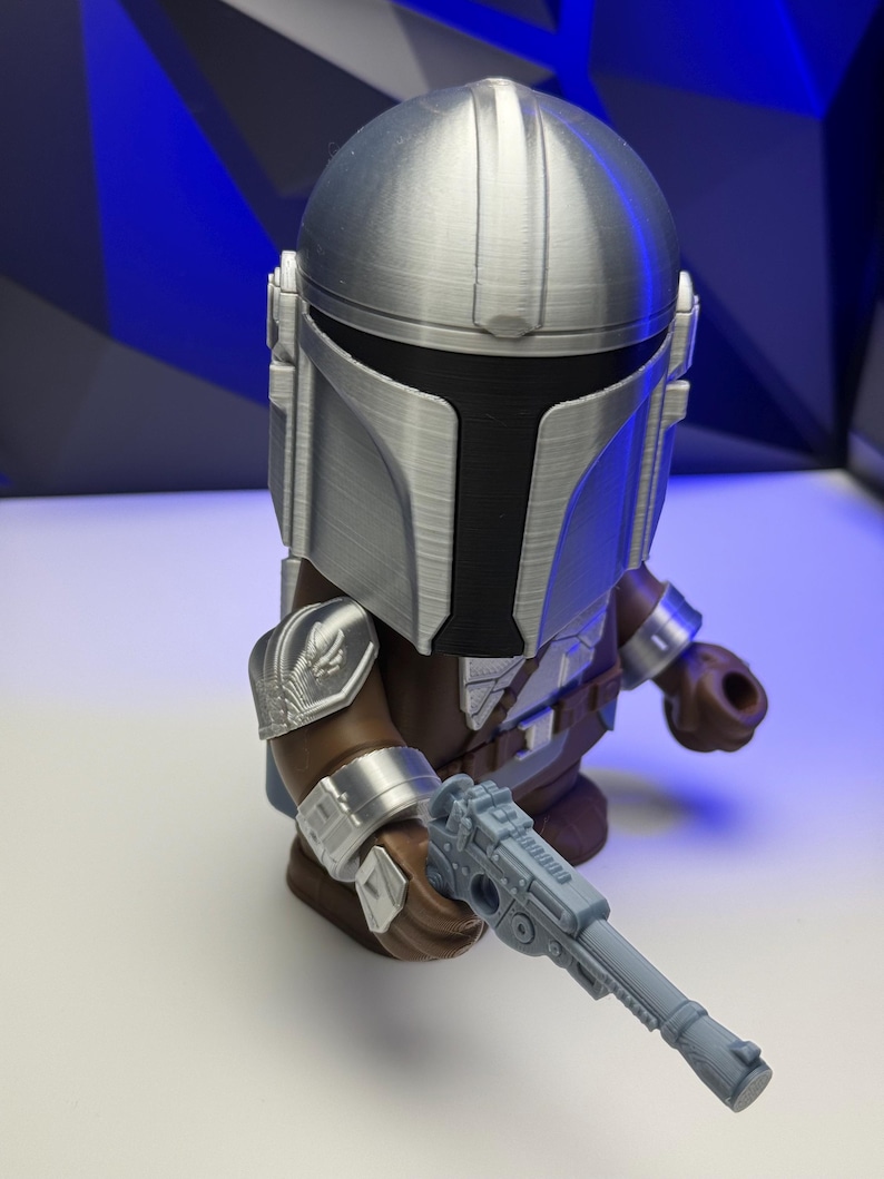 May include: A silver and black Mandalorian action figure, holding a gray blaster pistol. The figure has a detailed helmet and armor, with brown and silver accents. The background is a blue geometric pattern.