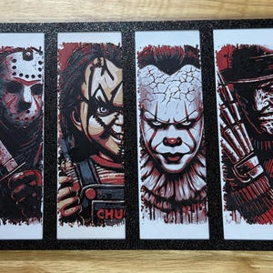 May include: A framed print featuring four iconic horror movie characters: Jason Voorhees, Chucky, Pennywise, and Freddy Krueger. Each character is rendered in red, black, and white, with a black border around each image. The overall theme is horror.