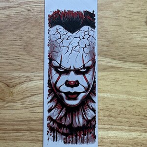 May include: A rectangular sticker depicting a detailed illustration of a menacing clown face. The clown has white face paint, red accents, and a cracked forehead design. The image is set against a light background.