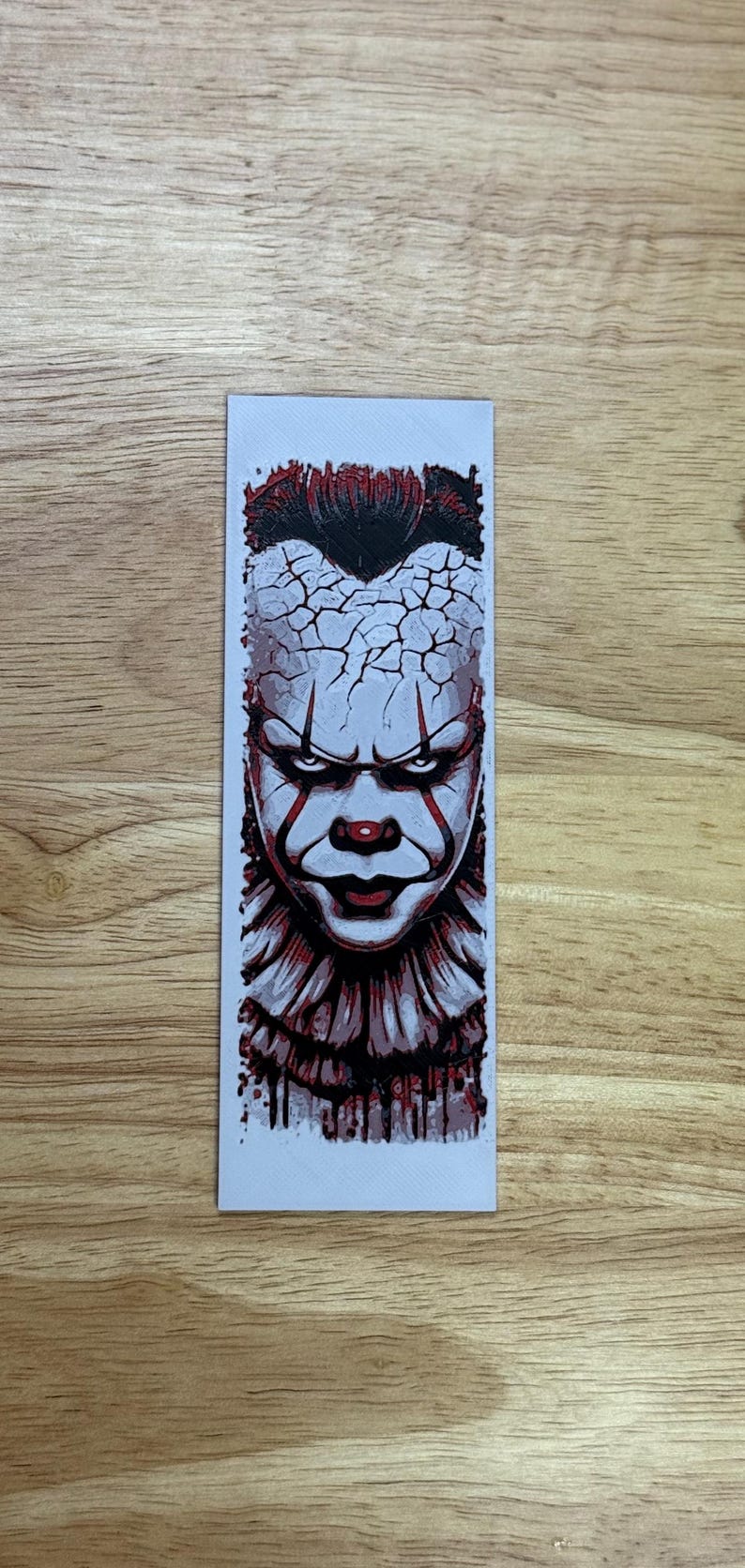 May include: A rectangular sticker depicting a detailed illustration of a menacing clown face. The clown has white face paint, red accents, and a cracked forehead design. The image is set against a light background.