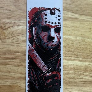 May include: A rectangular bookmark with a graphic illustration of a hockey mask-wearing figure holding a bloody knife. The design is mainly black and red, with a white background and a wooden surface.