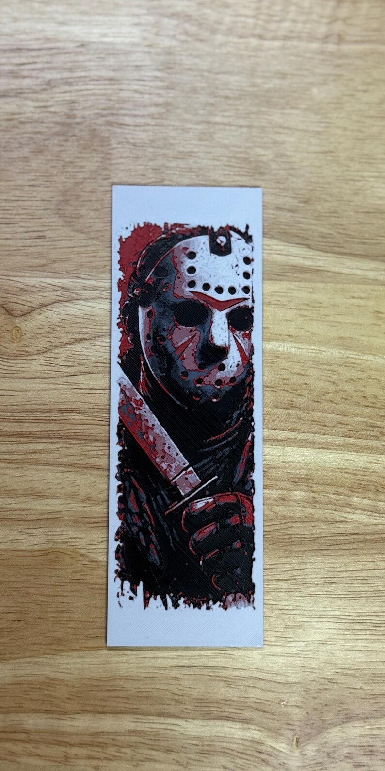 May include: A rectangular bookmark with a graphic illustration of a hockey mask-wearing figure holding a bloody knife. The design is mainly black and red, with a white background and a wooden surface.