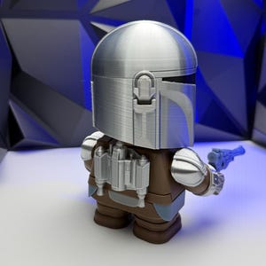 May include: A silver-colored Mandalorian figure with a helmet, brown armor, and a blue blaster. The figure has a detailed design and is standing on a white surface. The background features a geometric pattern.