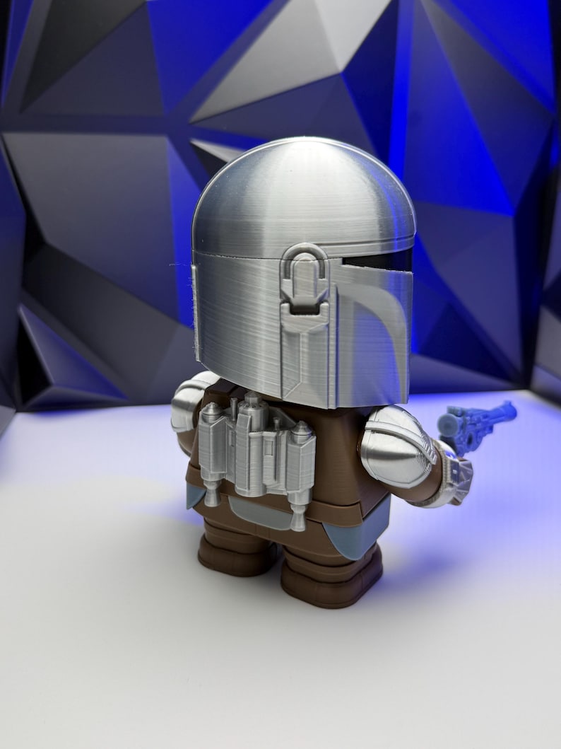 May include: A silver-colored Mandalorian figure with a helmet, brown armor, and a blue blaster. The figure has a detailed design and is standing on a white surface. The background features a geometric pattern.