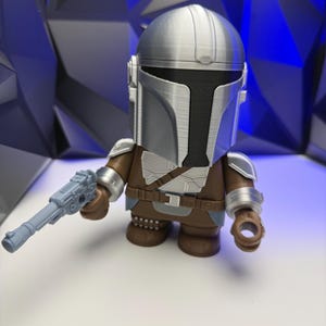 May include: A stylized figure of The Mandalorian, featuring a silver helmet and armor, brown clothing, and a gray blaster. The figure is posed against a blue and black geometric background.