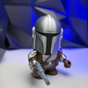 May include: A silver and brown Mandalorian figure with a blaster. The figure has a detailed helmet and armor, and is holding a light blue blaster. The figure is set against a blue geometric background.