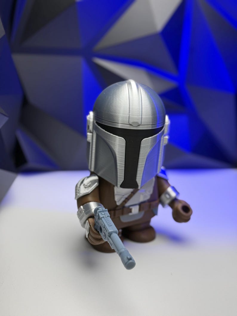 May include: A silver and brown Mandalorian figure with a blaster. The figure has a detailed helmet and armor, and is holding a light blue blaster. The figure is set against a blue geometric background.