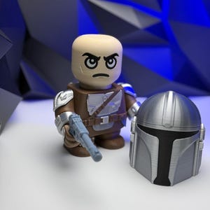 May include: A 3D-printed action figure of a character with a stern expression, wearing brown armor, a silver pauldron, and holding a light blue blaster. A silver helmet is placed next to the figure. The background is a geometric blue design.