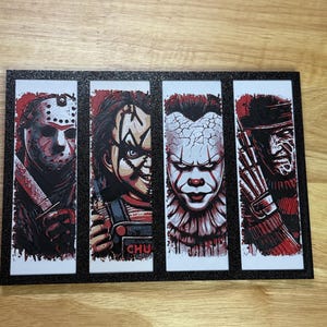 May include: A framed art print featuring four horror movie characters: Jason Voorhees, Chucky, Pennywise, and Freddy Krueger. Each character is depicted in a vertical panel with a red and white colour scheme. The frame is black with glitter.