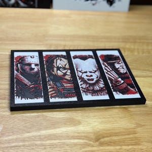 May include: A black rectangular decorative item featuring four horror movie characters: Jason Voorhees, Chucky, Pennywise, and Freddy Krueger. Each character is depicted in a vertical panel with a red and black colour scheme. The item is displayed on a wooden surface.
