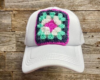 May include: White trucker hat with a crocheted granny square patch in pink, green, and gray.