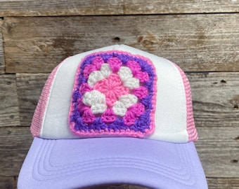 May include: A white trucker hat with a purple, pink, and white crocheted square patch on the front. The hat has a light purple brim.