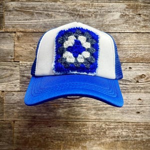 May include: A trucker hat with a white front panel and a blue mesh back. The hat features a blue crocheted square with a white and blue floral design. The brim is bright blue.