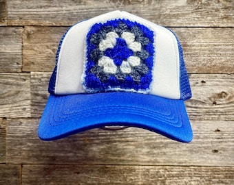 May include: A trucker hat with a white front panel and a blue mesh back. The hat features a blue crocheted square with a white and blue floral design. The brim is bright blue.