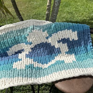 May include: A chunky knit blanket with a blue and white color gradient pattern. The blanket is made with thick yarn and has a soft, fluffy texture.