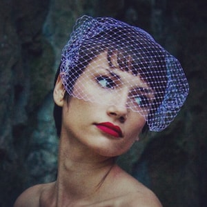 May include: A woman wearing a white veil with a fine mesh pattern. The veil is covering her forehead and eyes. She is wearing red lipstick and has a neutral expression.