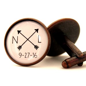 May include: A pair of cufflinks with a compass design featuring the initials "N" and "L" and the date "9-27-16". The cufflinks are made of copper and have a white background.