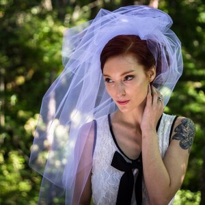 Puede incluir: Una mujer con un velo blanco y un vestido blanco con un lazo negro. Tiene el pelo rojo y est&aacute; de pie en un bosque.