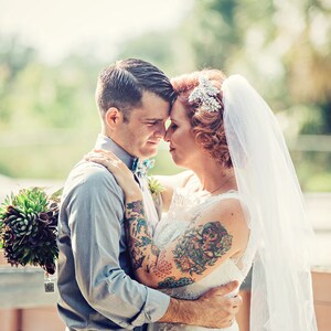 Puede incluir: Una novia y un novio con tatuajes se abrazan. La novia lleva un vestido de novia blanco con velo y una diadema de joyas. El novio lleva una camisa de botones azul claro y pantalones oscuros. Est&aacute;n de pie al aire libre con vegetaci&oacute;n en el fondo.