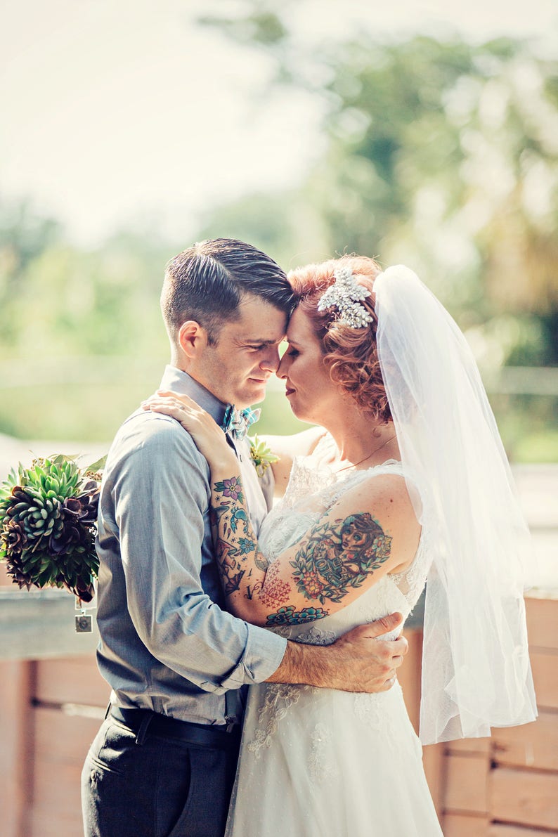 Puede incluir: Una novia y un novio con tatuajes se abrazan. La novia lleva un vestido de novia blanco con velo y una diadema de joyas. El novio lleva una camisa de botones azul claro y pantalones oscuros. Est&aacute;n de pie al aire libre con vegetaci&oacute;n en el fondo.