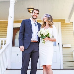 May include: A couple poses for a wedding photo on a porch with white railings. The groom is wearing a dark blue suit and the bride is wearing a white dress with a veil. They are holding a bouquet of white flowers.