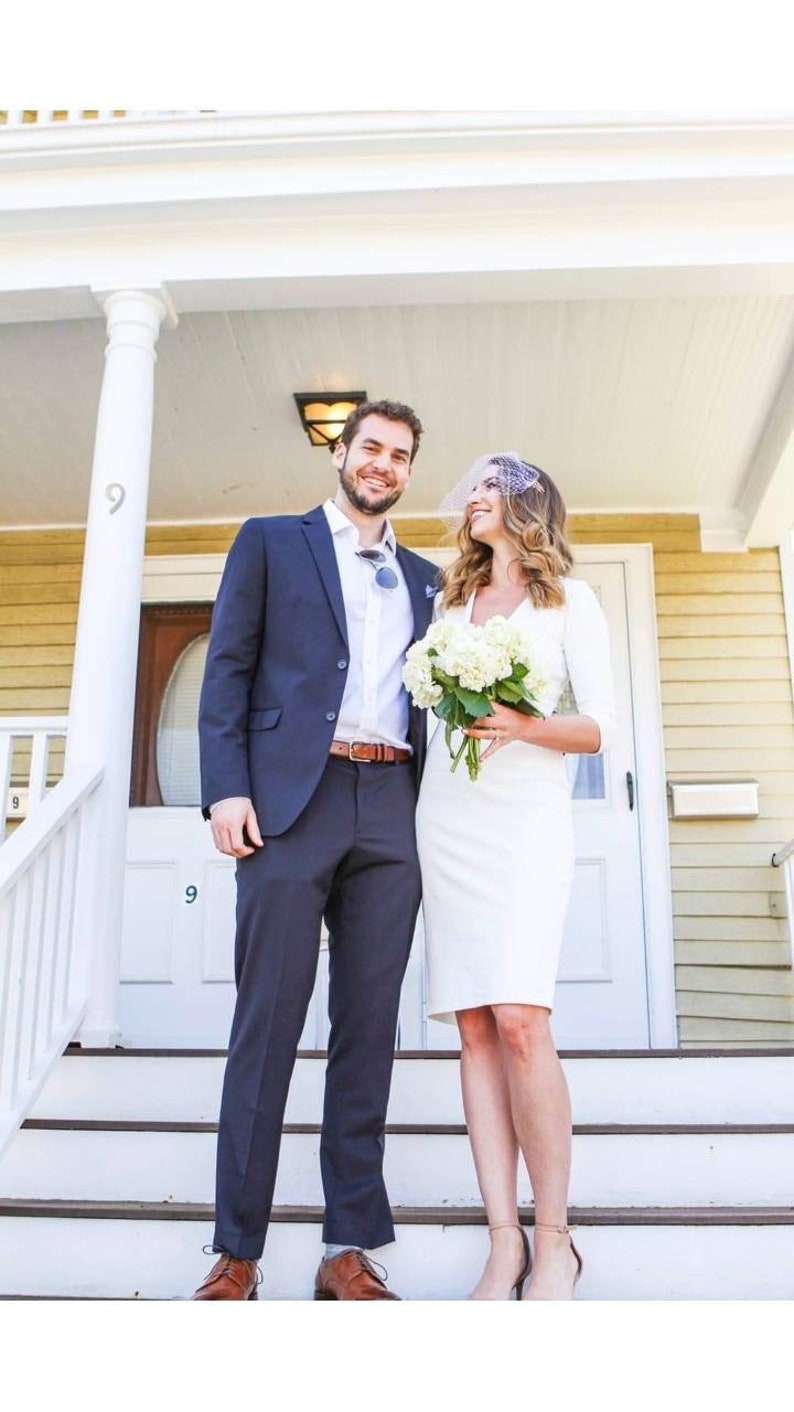 May include: A couple poses for a wedding photo on a porch with white railings. The groom is wearing a dark blue suit and the bride is wearing a white dress with a veil. They are holding a bouquet of white flowers.
