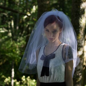 Puede incluir: Una mujer con un velo blanco y un vestido blanco con un lazo negro. Est&aacute; de pie en un bosque.