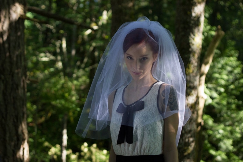 Puede incluir: Una mujer con un velo blanco y un vestido blanco con un lazo negro. Est&aacute; de pie en un bosque.