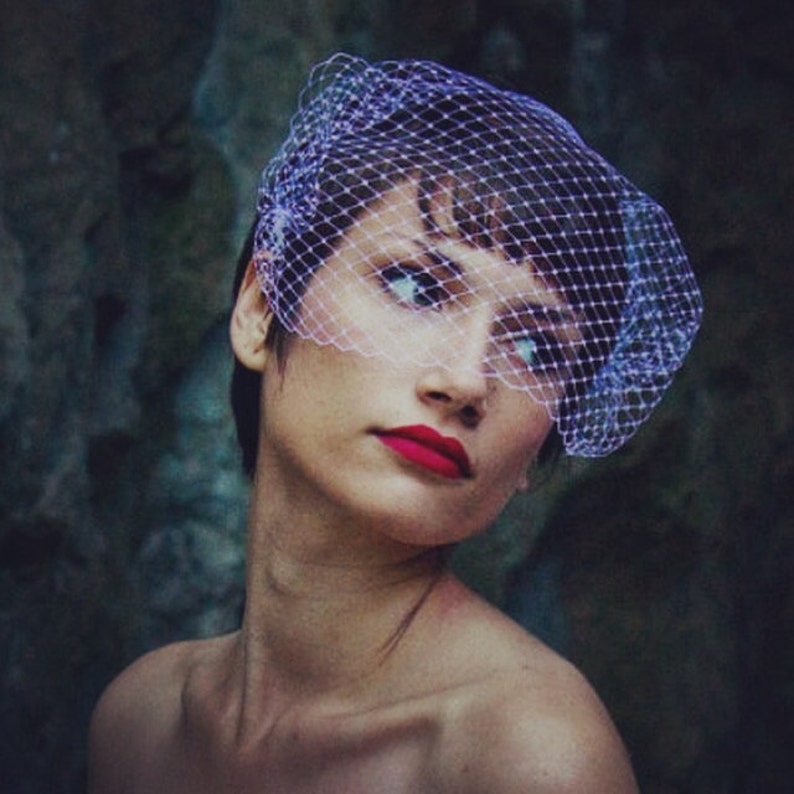 May include: A woman wearing a white veil with a fine mesh pattern. The veil is covering her forehead and eyes. She is wearing red lipstick and has a neutral expression.