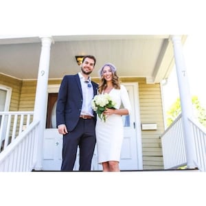 May include: A couple poses for a wedding photo on a porch with white railings. The groom is wearing a dark blue suit and the bride is wearing a white dress with a white flower bouquet.
