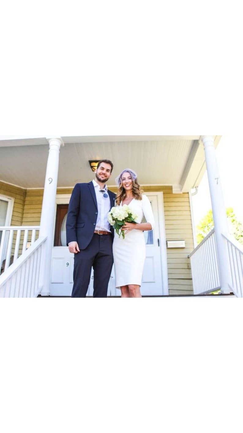May include: A couple poses for a wedding photo on a porch with white railings. The groom is wearing a dark blue suit and the bride is wearing a white dress with a white flower bouquet.