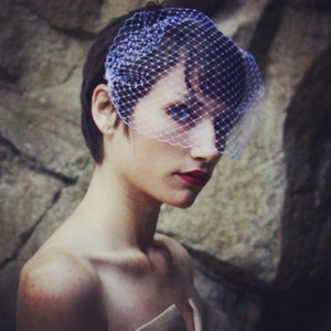 May include: A woman wearing a white veil with a fine mesh pattern. The veil is covering her hair and part of her face. She is wearing red lipstick.