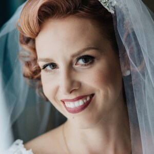 Puede incluir: Una novia con el pelo rojo que lleva un velo blanco con una diadema de joyas. Est&aacute; sonriendo y mirando a la c&aacute;mara. Lleva un vestido blanco de encaje.
