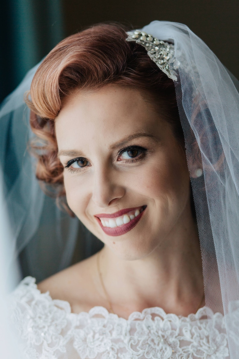 Puede incluir: Una novia con el pelo rojo que lleva un velo blanco con una diadema de joyas. Est&aacute; sonriendo y mirando a la c&aacute;mara. Lleva un vestido blanco de encaje.