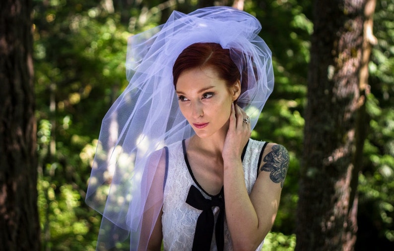 Puede incluir: Una mujer con un velo blanco y un vestido blanco con un lazo negro. Tiene el pelo rojo y est&aacute; de pie en un bosque.